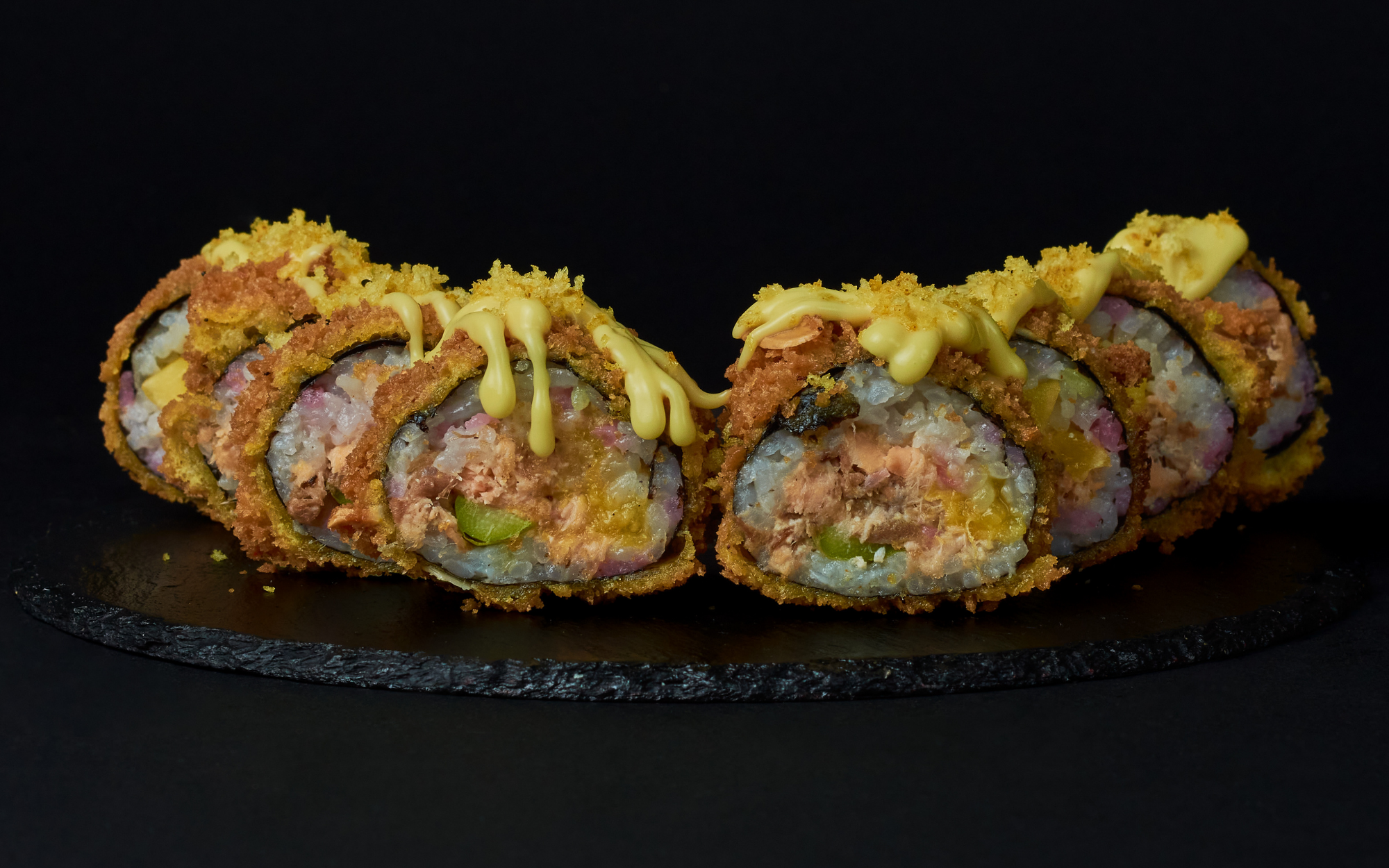 Sechs frittierte Sushi-Rollen, gefüllt mit Fisch und Gemüse, überzogen mit gelber Soße und knusprigen Bröseln, sind in einer Reihe auf einer schwarzen Schieferplatte vor dunklem Hintergrund angeordnet.