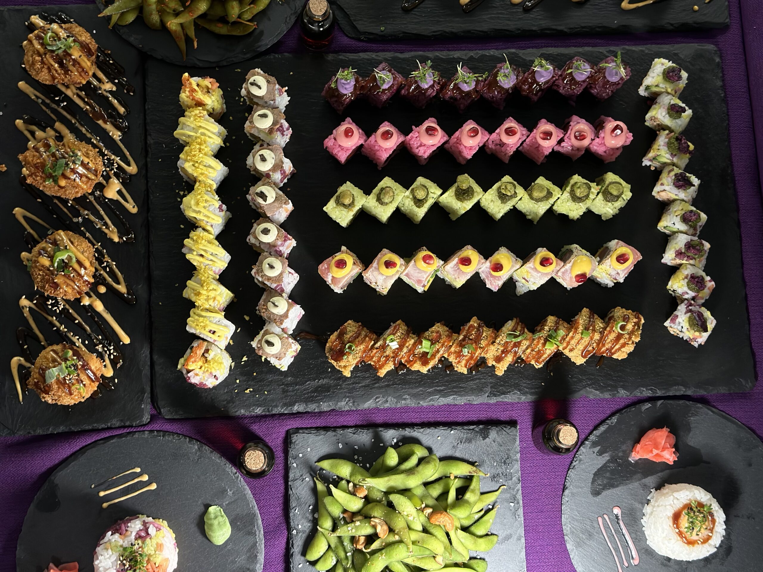 Ein Blick von oben auf verschiedene Sushi-Rollen, Edamame und Beilagen, die ordentlich auf schwarzen Schieferplatten über einem lila Tischtuch angeordnet sind. Das Sushi ist bunt, mit verschiedenen Toppings und Saucen.