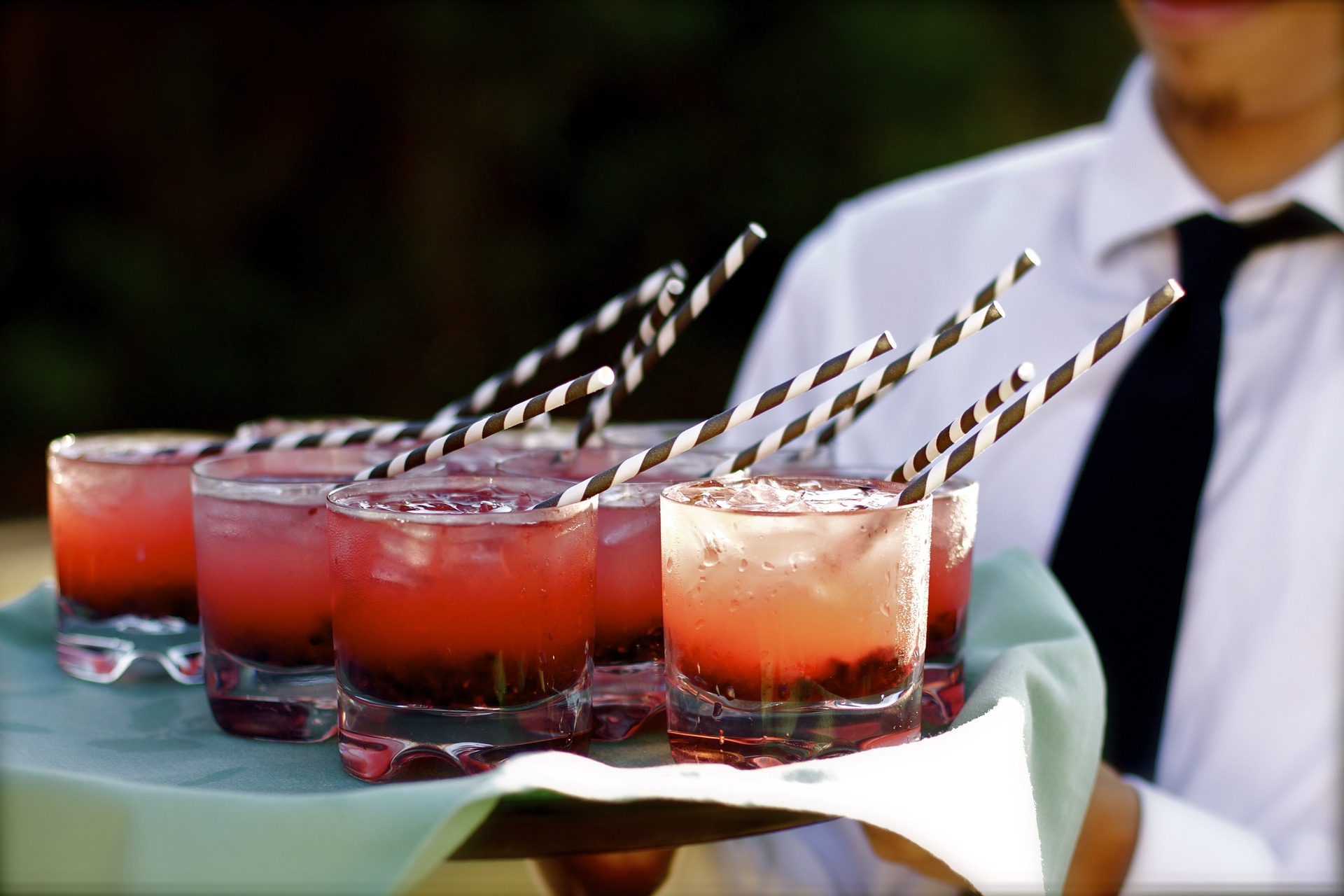 Ein Kellner in weißem Hemd und schwarzer Krawatte hält ein Tablett mit rosa Cocktails mit gestreiften Papierstrohhalmen, jedes Glas mit Eis gefüllt und mit Beeren garniert, vor einem unscharfen Hintergrund im Freien.