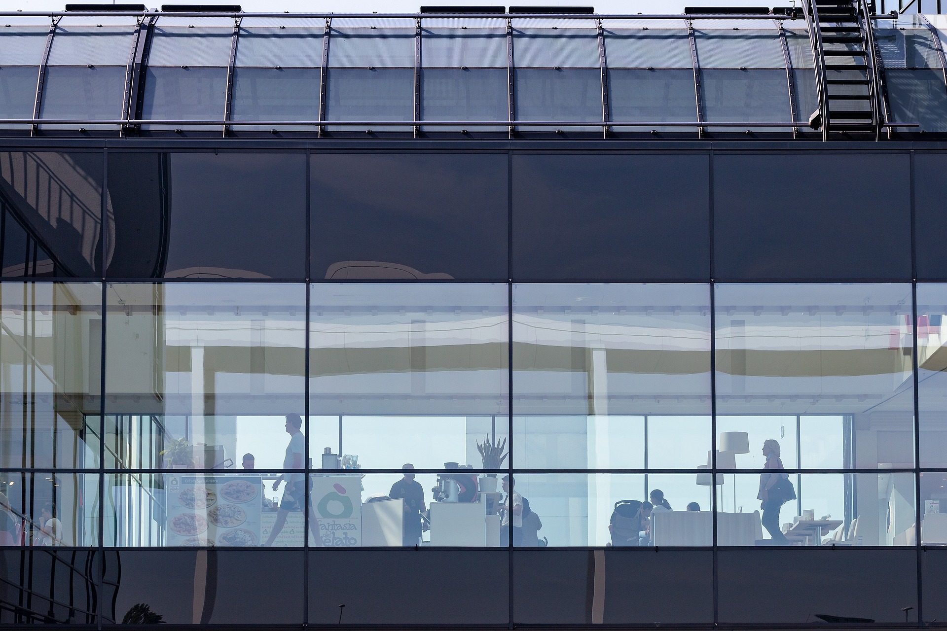 Menschen sitzen und stehen in einem modernen Gebäude mit Glaswänden, die durch große Fenster sichtbar sind. Die Szene zeigt einen geräumigen, gut beleuchteten Innenraum mit Tischen, Stühlen und ein paar dekorativen Pflanzen.