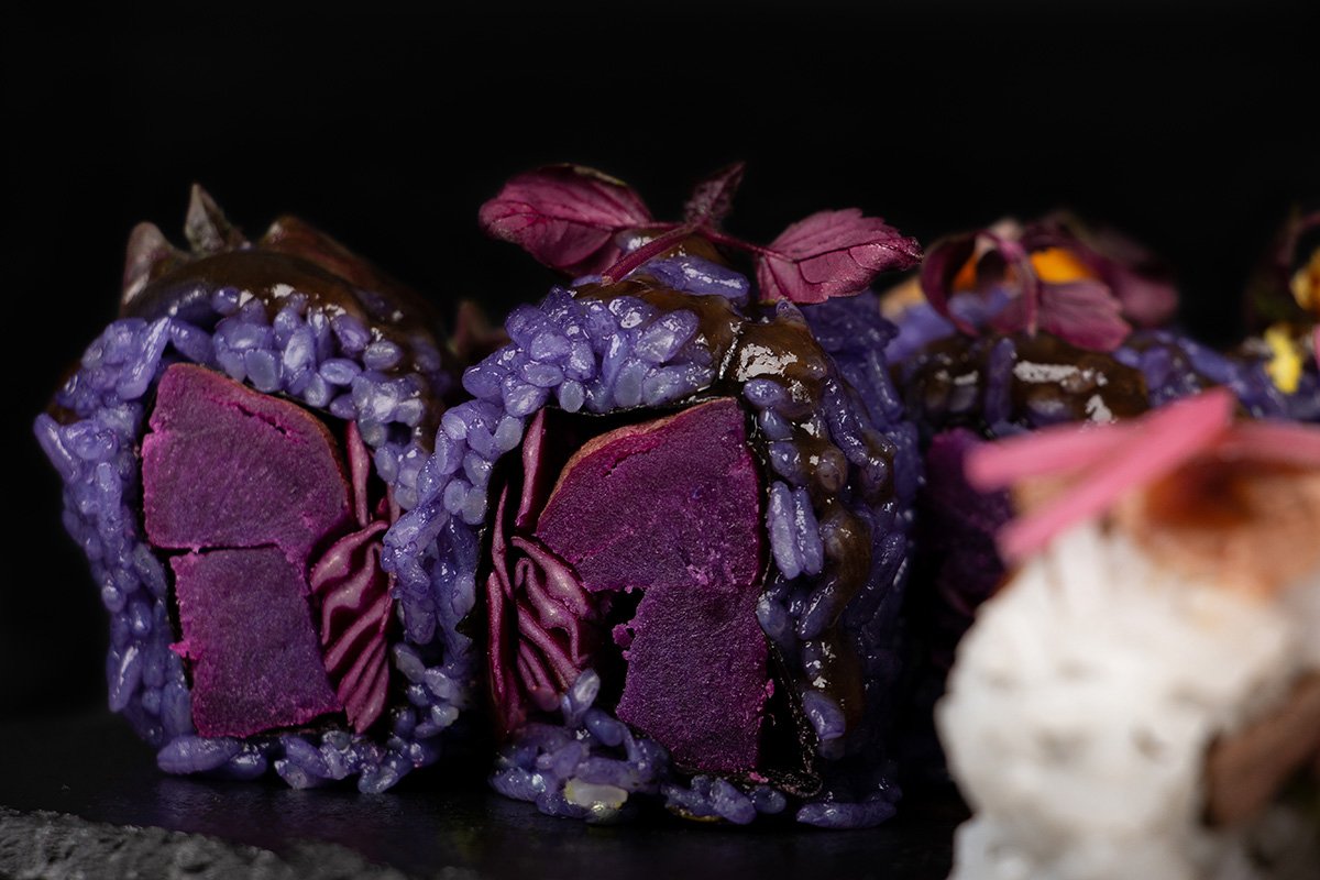 Nahaufnahme von Sushi-Rollen mit leuchtend violettem Reis und violettem Gemüse, garniert mit kleinen violetten Blättern, vor einem dunklen Hintergrund. Die Farben verleihen den Sushi ein einzigartiges und auffälliges Aussehen.