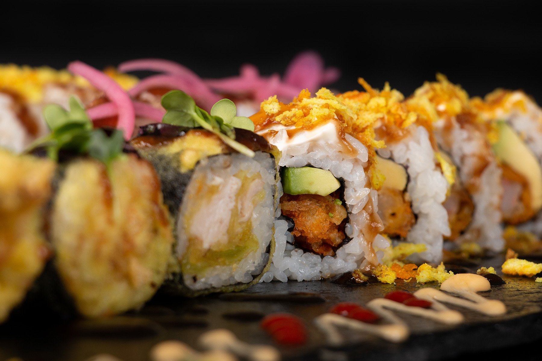 Nahaufnahme von verschiedenen Sushi-Rollen, die mit Saucen, knusprigen Krümeln und Garnierungen wie eingelegten Zwiebeln und Mikrogrün auf einem schwarzen Schieferbrett angerichtet sind.