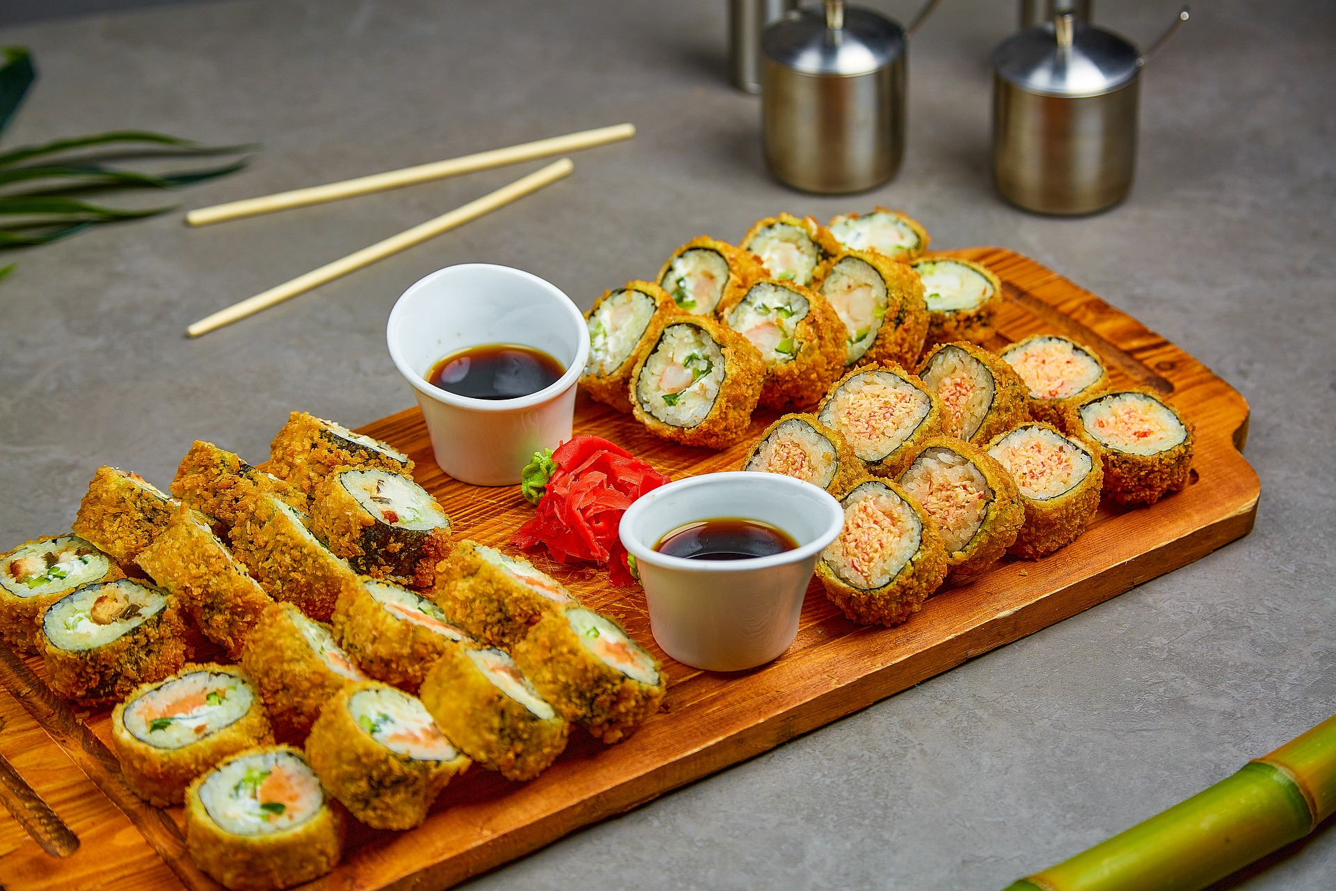 Ein Holzbrett mit verschiedenen gebratenen Sushi-Rollen, zwei kleinen Bechern mit Sojasauce, eingelegtem Ingwer und Essstäbchen steht auf einem grauen Tisch.