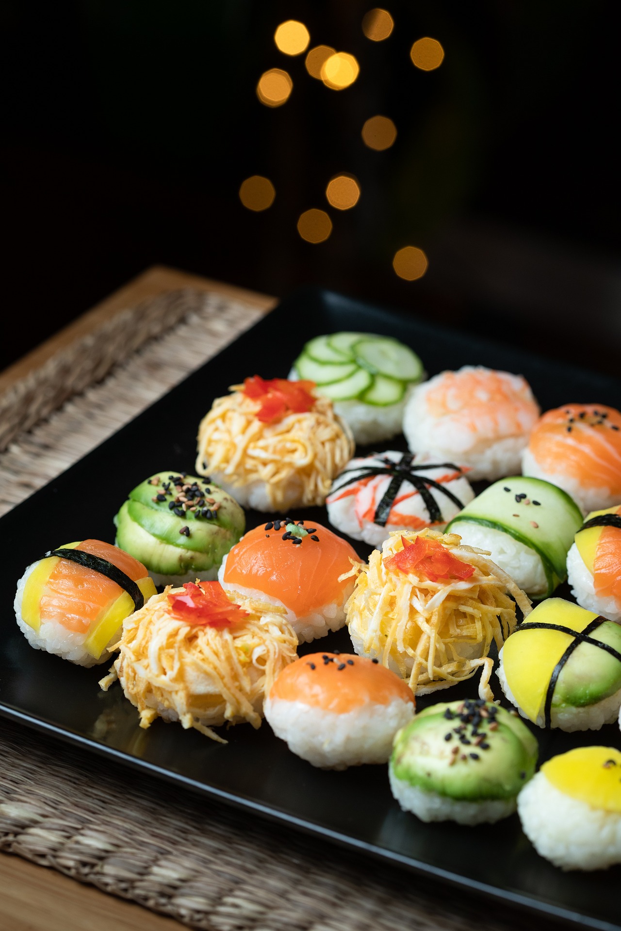 Auf einem schwarzen Teller liegen verschiedene Sushi-Bällchen mit Gurkenscheiben, geraspeltem Ei, Lachs, Mango und Avocado, garniert mit Nori-Streifen, Sesamkörnern und kleinen roten Belägen. Im Hintergrund flackern warme Lichterketten.